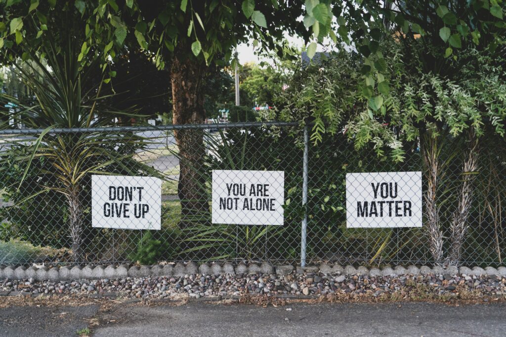 Overcoming REcovery Challenges is shown in a chain-link fence adorned with three motivational signs in a leafy suburban area. The signs, displayed in sequence, read "DON'T GIVE UP," "YOU ARE NOT ALONE," and "YOU MATTER," in bold, black lettering on a white background. The backdrop features an assortment of trees and plants partially obscuring the fence, under a clear daylight sky, suggesting a message of hope and support for individuals facing recovery challenges.