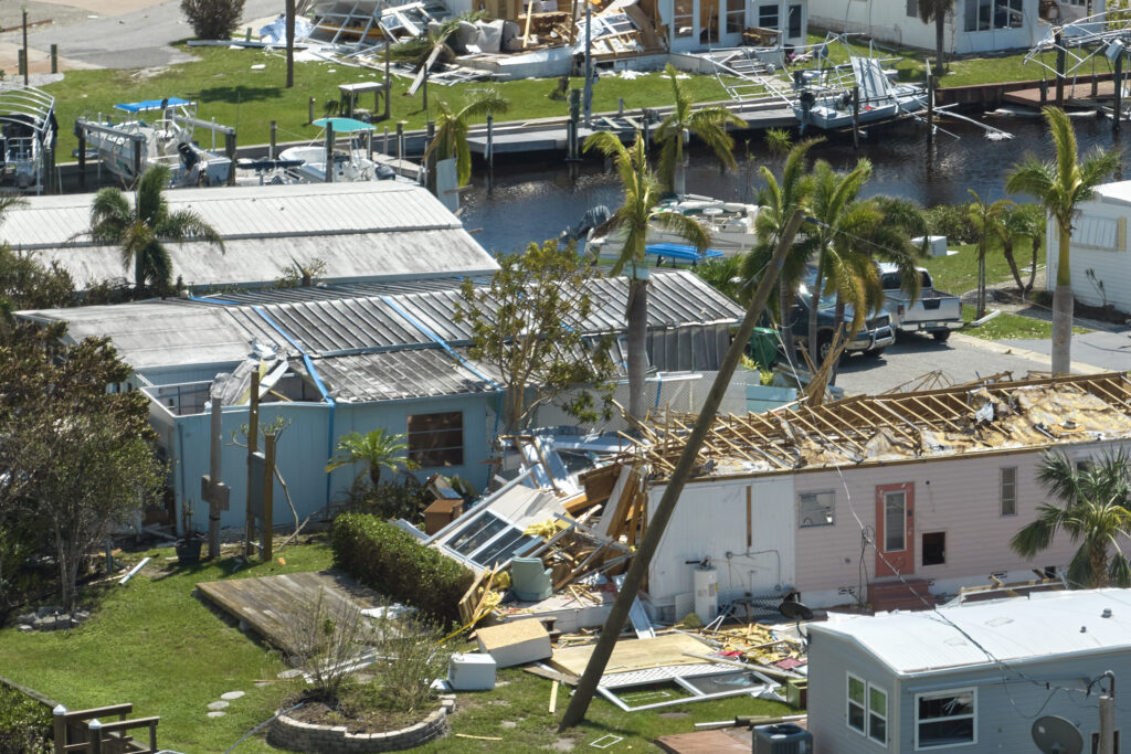 An aerial view of residential homes damaged by a hurricane, with roofs partially torn off, debris scattered across the lawns, and boats docked near the waterfront. Palm trees are bent, and structures appear severely impacted, indicating the aftermath of a natural disaster.