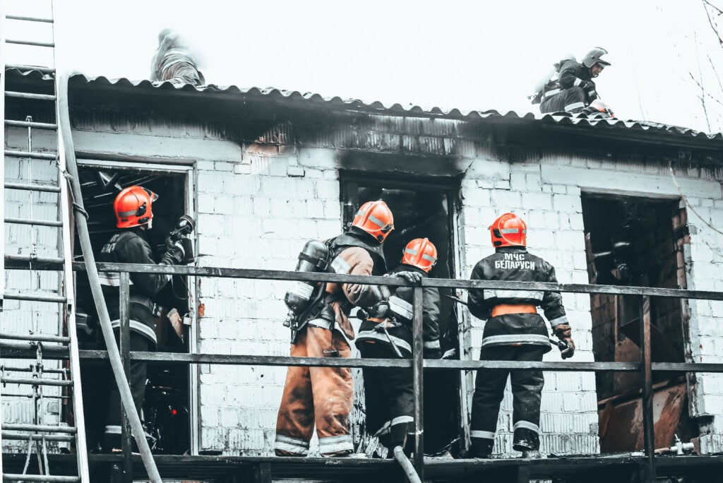 Salvaging equipmnents and inventory, the Firefighters in full gear are actively extinguishing a fire in a brick building. The image shows several firefighters, some positioned on scaffolding and one emerging from a smoky window, while another firefighter is seen on the roof. Their helmets have "MЧC БЕЛАРУСЬ" printed on them, indicating they are members of the Ministry of Emergency Situations of Belarus. The building appears damaged with visible soot and smoke, highlighting the intensity of the firefighting efforts.