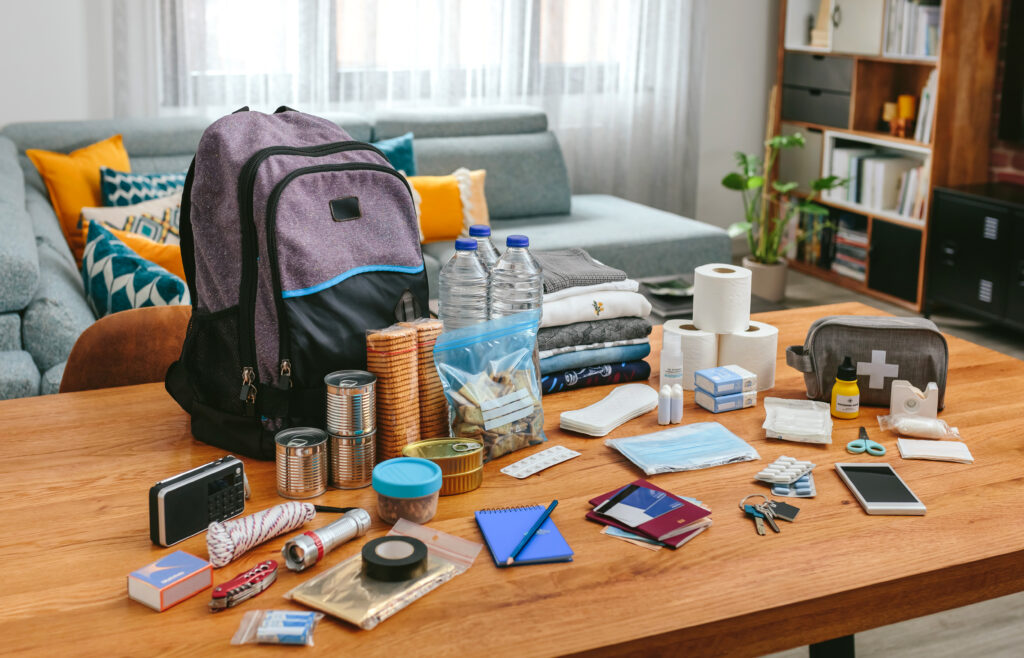 Creating a disaster preparedness budget starts with an organized display of emergency preparedness items on a table, including a backpack, canned food, bottled water, first aid kit, flashlight, and essential documents, ready for disaster preparedness.
