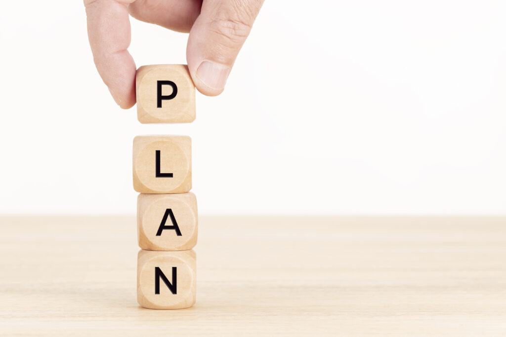 A hand arranging wooden blocks vertically, each block containing a letter to form the word 'PLAN.' This image symbolizes strategic organization and the importance of planning. Regular updates to your disaster plan are crucial to ensure it reflects current risks, resources, and best practices, helping businesses and individuals remain resilient and prepared for unforeseen emergencies.