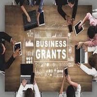 Overhead view of a group of professionals working together at a table with laptops and tablets, with the words 'Business Grants' displayed in the center, representing discussions or planning for securing disaster recovery grants.